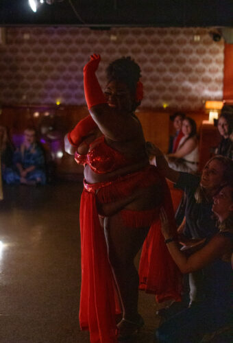 burlesque performer in red costume dances for audience members as they tip her